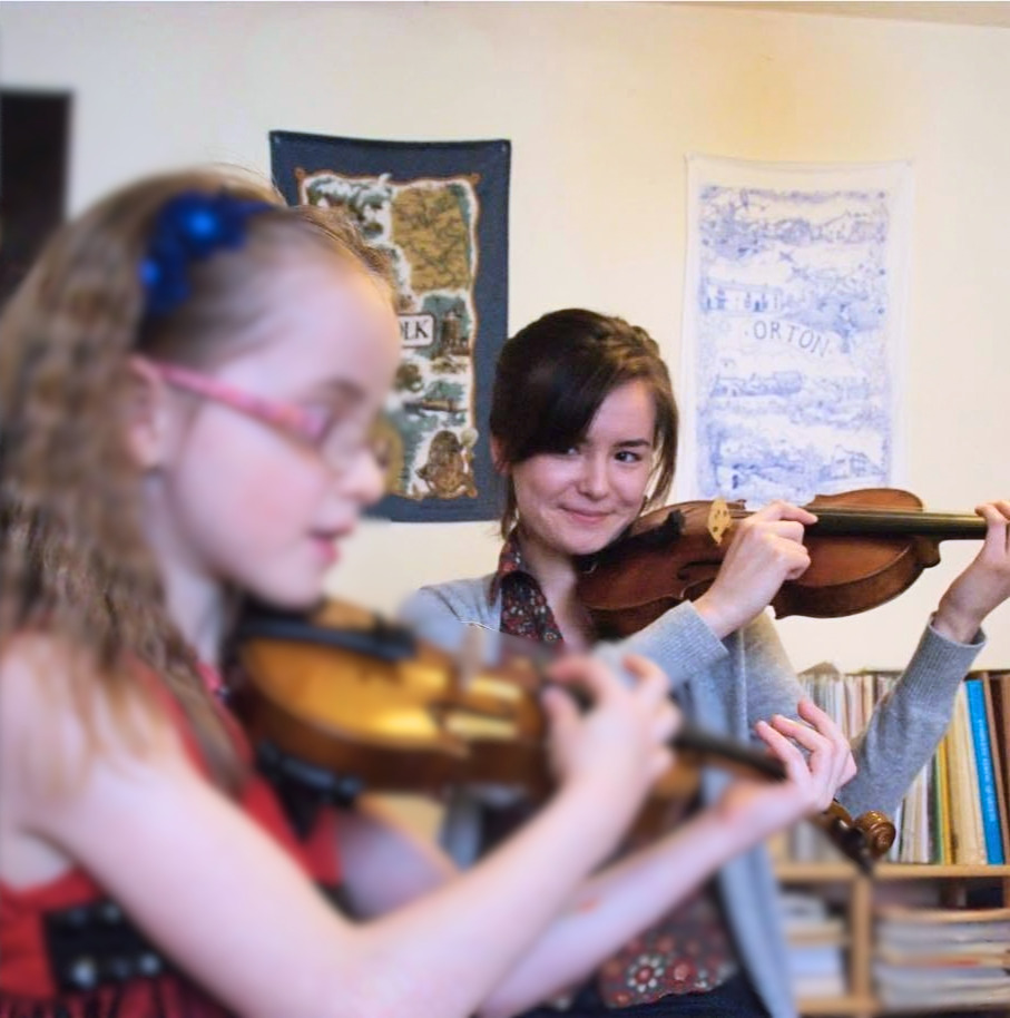 Violin teacher smiling as she plays alongside a young student during a lesson